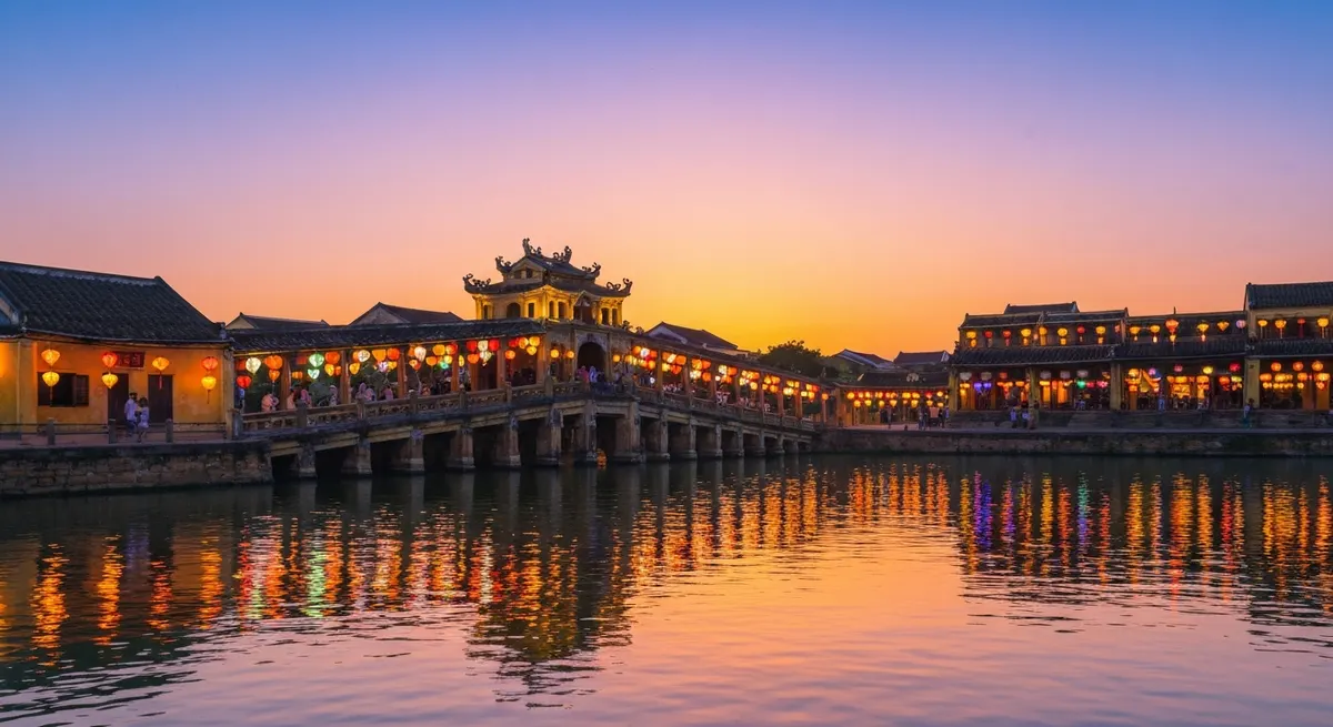 黄昏时分从怀河水边看会安日本廊桥，红黄灯笼的光芒在水面上闪烁，桥上行人身影绰绰